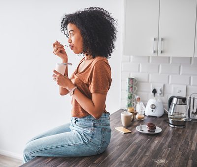 Shot of a young woman having a tub of yoghurt in the kitchen at home