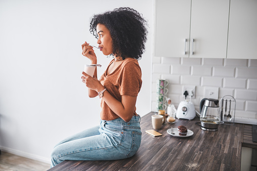Shot of a young woman having a tub of yoghurt in the kitchen at home